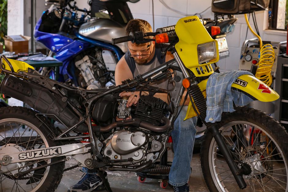 Mechanic repairing a Suzuki motorcycle in a cluttered garage, focusing on engine maintenance.