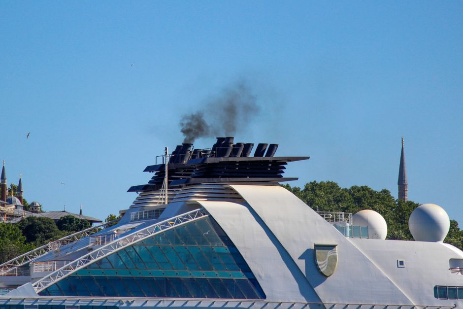 Cruise ship chimney emitting smoke under a clear sky with a cityscape backdrop.