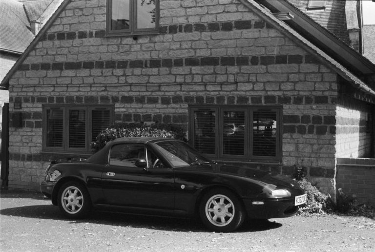 Black and white photo of a classic Mazda MX-5 Miata parked by a rustic stone house.