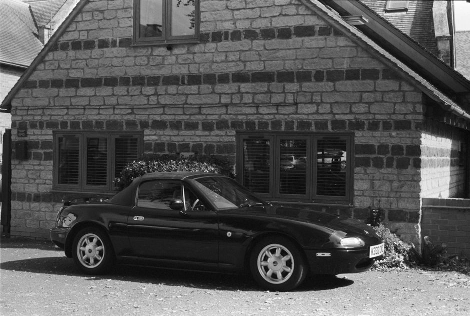 Black and white photo of a classic Mazda MX-5 Miata parked by a rustic stone house.