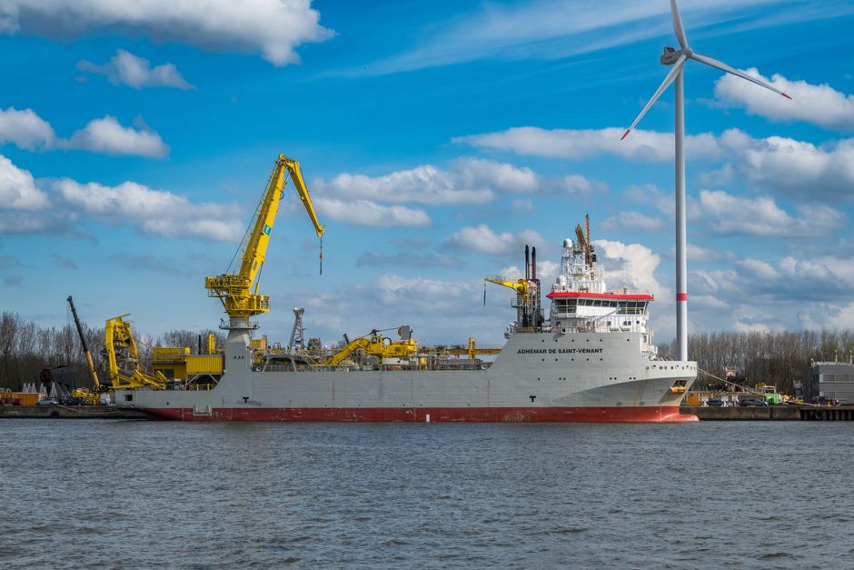 A pipe burying vessel moored at a harbor with a wind turbine nearby.