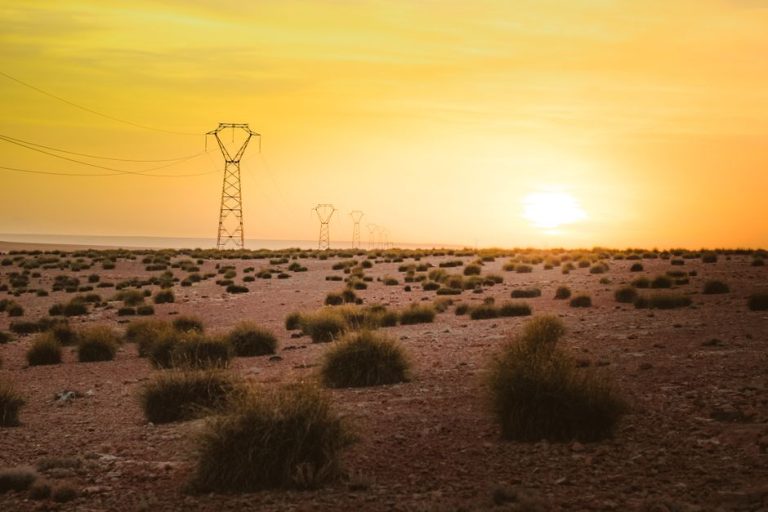 Power lines stretch across an arid desert landscape amidst a glowing sunset.