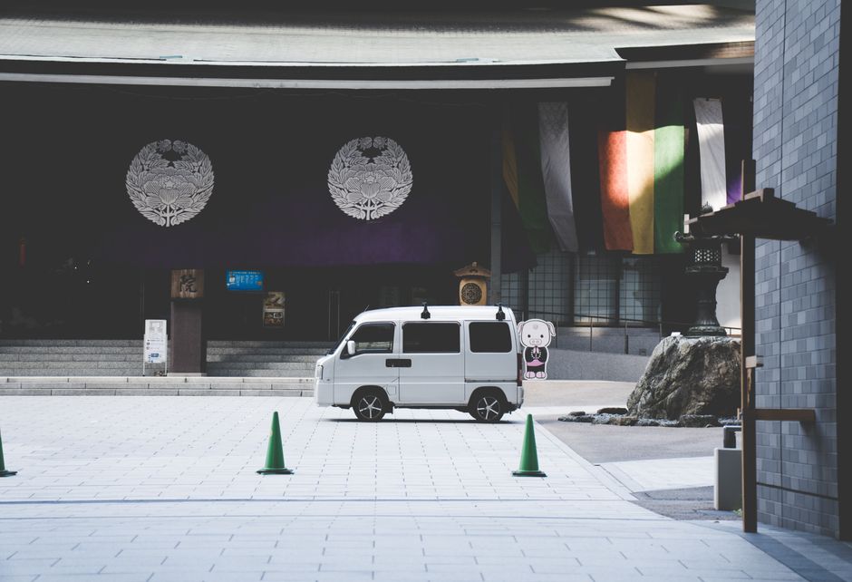 White kei van parked in front of an Osaka shrine with colorful banners.