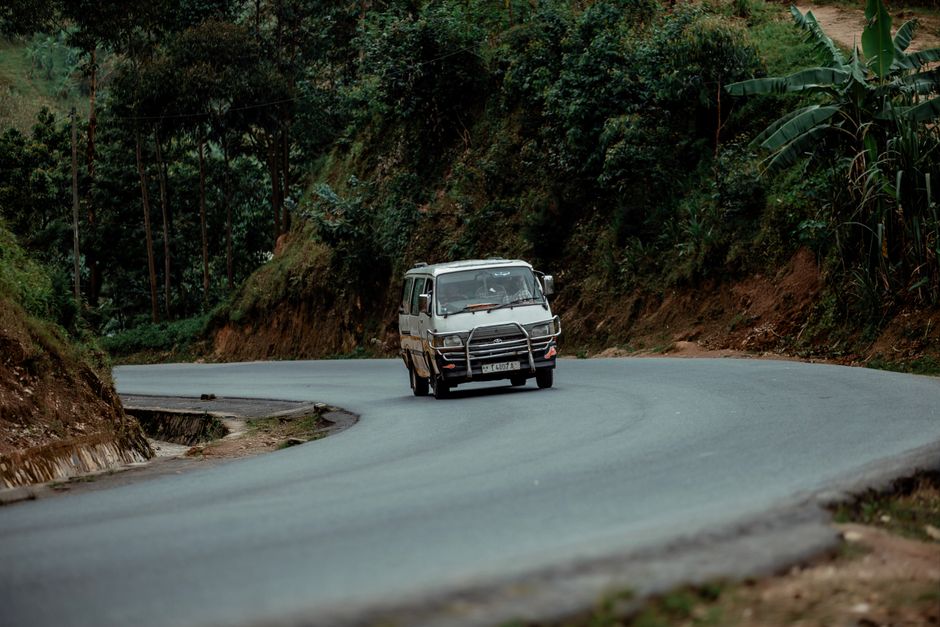 A white minibus drives through a lush, winding rural road surrounded by greenery.