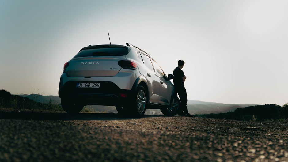 A man leans against a Dacia car on a scenic road at sunset, capturing a sense of wanderlust.