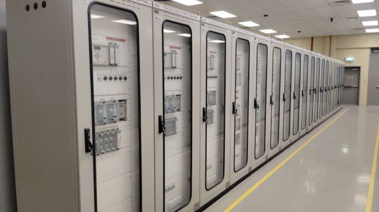 View of an industrial control room featuring rows of electrical panels in a well-lit environment.