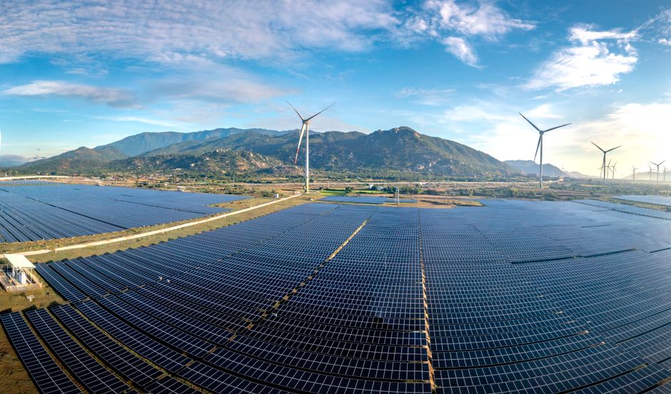 Aerial view of a vast solar farm with wind turbines against a mountainous backdrop on a sunny day.
