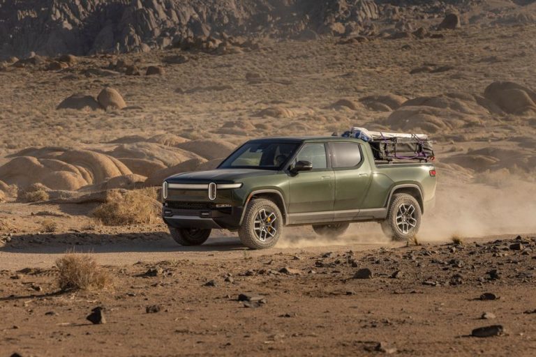 An electric pickup truck navigates a rocky desert trail, raising dust in a rugged landscape.