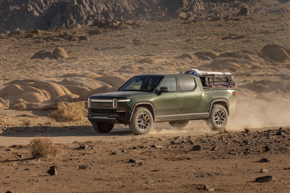 An electric pickup truck navigates a rocky desert trail, raising dust in a rugged landscape.