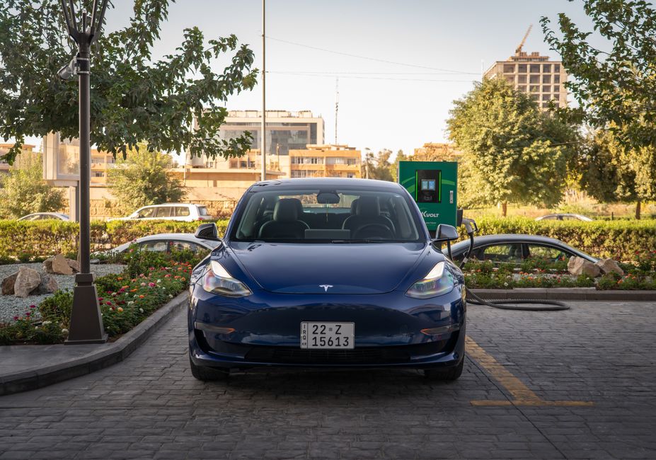 A blue Tesla Model 3 charges at an outdoor station during the day, surrounded by greenery and urban architecture.