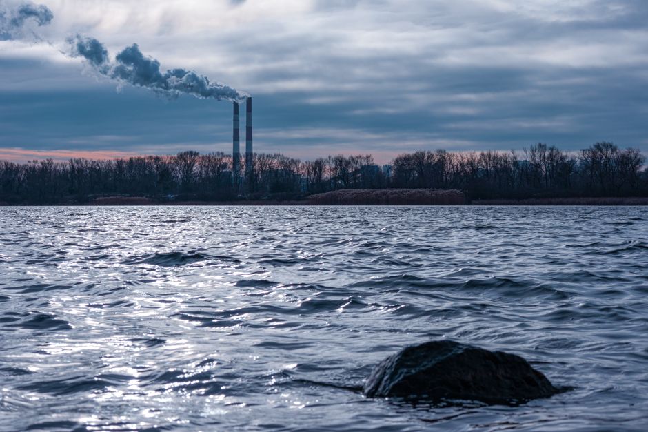 Factory smoke stacks emitting pollution over river with dramatic sky in Kyiv.
