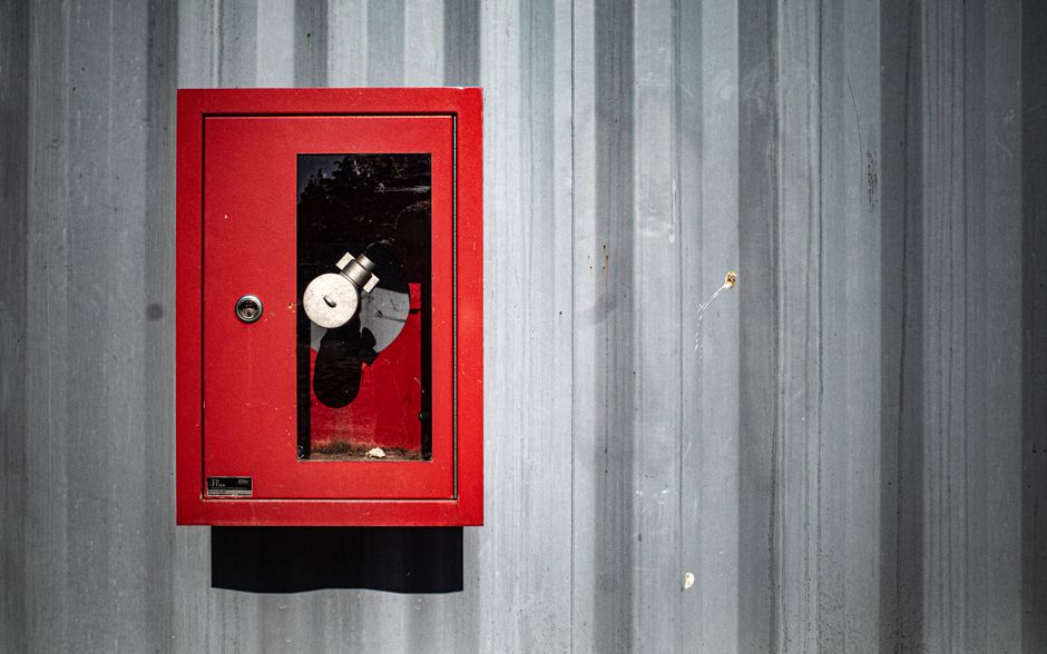 Bold red fire hose cabinet on industrial corrugated wall under bright sunlight.