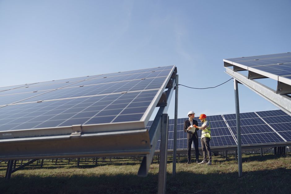 Supervisor in hardhat and formal wear discussing project with workman in uniform while standing with papers near modern solar panels under blue sky