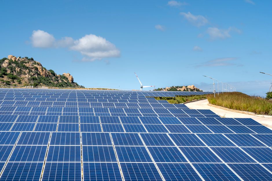 Expansive solar farm with rows of panels under clear blue sky and hills in the background.