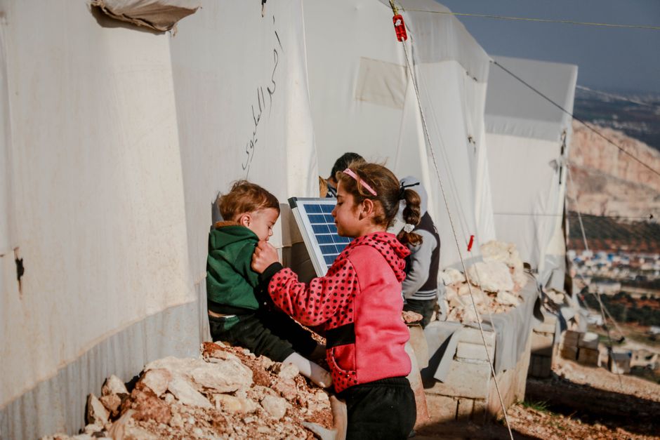 Side view of ethnic girl taking care of brother sitting on stone rubble outside tents in refugee camp