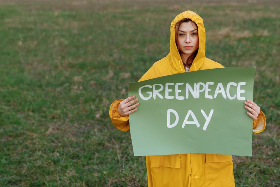 A woman in a yellow raincoat holds a 'Greenpeace Day' sign on a grassy field.