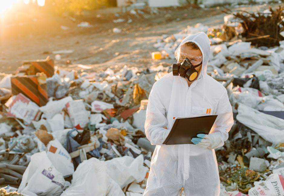 Scientist in protective gear documenting pollution at a garbage dump site outdoors.