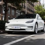 A sleek white Tesla Model 3 parked on a city street under trees, showcasing modern automotive design.