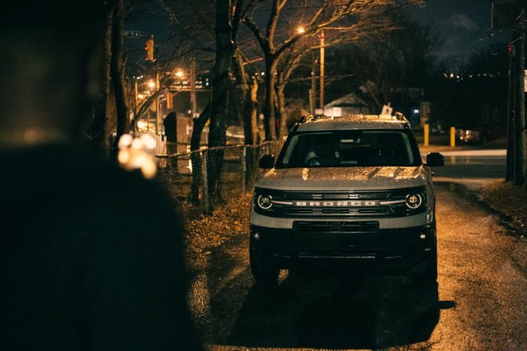 A white SUV parked on a wet urban street at night, surrounded by dim streetlights and trees.