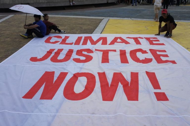 Large protest banner reading 'Climate Justice Now!' displayed outdoors during a demonstration.