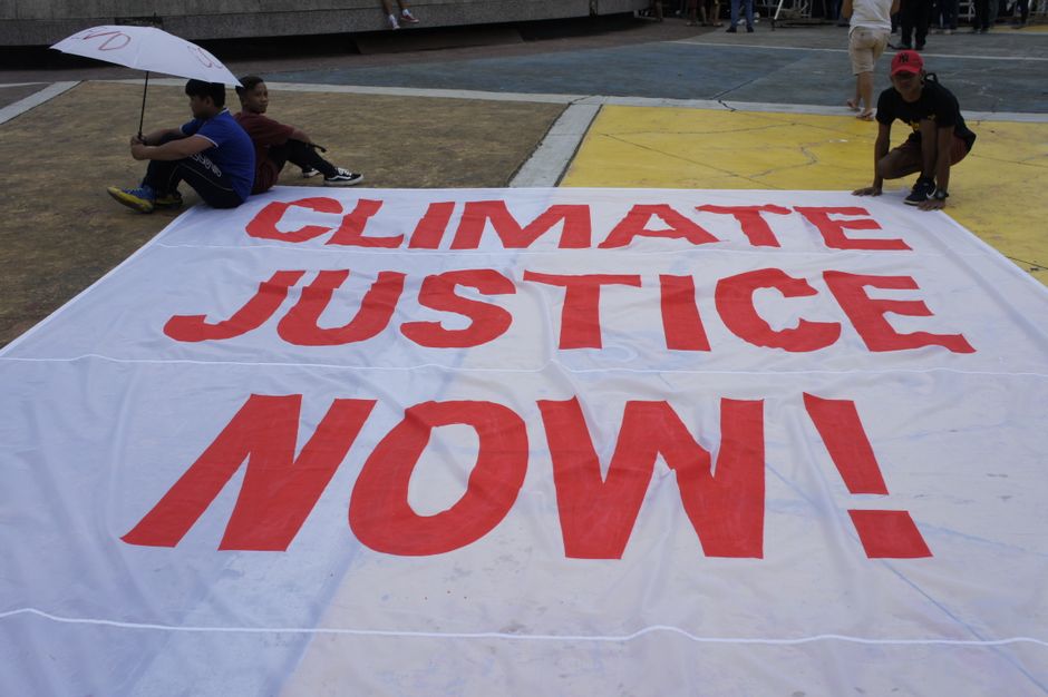 Large protest banner reading 'Climate Justice Now!' displayed outdoors during a demonstration.