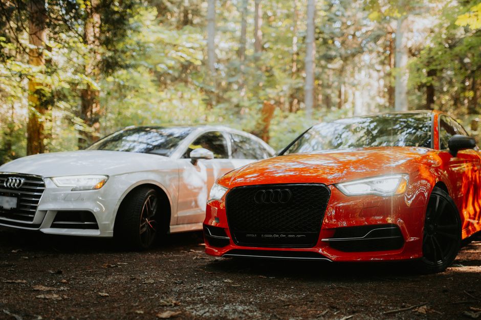Two modern Audi cars, red and white, parked in a serene forest setting.