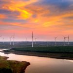 Aerial view of wind turbines in a vibrant sunset sky over a river and fields.