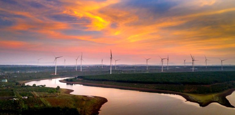 Aerial view of wind turbines in a vibrant sunset sky over a river and fields.