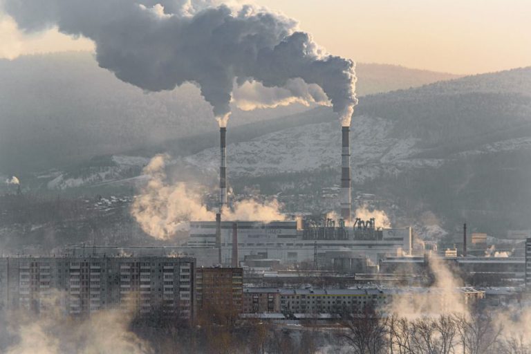 An industrial factory with chimneys emitting smoke, surrounded by snowy landscapes and buildings.