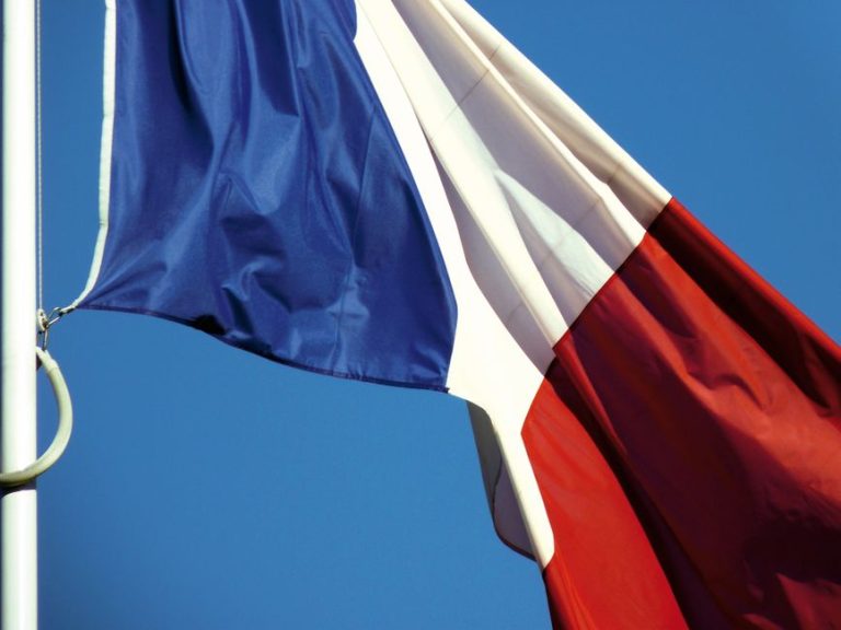 A close-up of the French national flag waving against a clear blue sky in Paris, France.