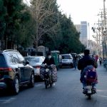 A bustling Beijing street with cars, scooters, and people, capturing urban life in daylight.