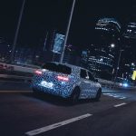 Electric vehicle with camo wrap driving through illuminated city at night, showcasing futuristic car design.