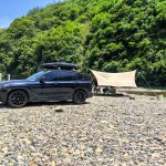 A black SUV parked beside a scenic river with a camping setup under a tent.