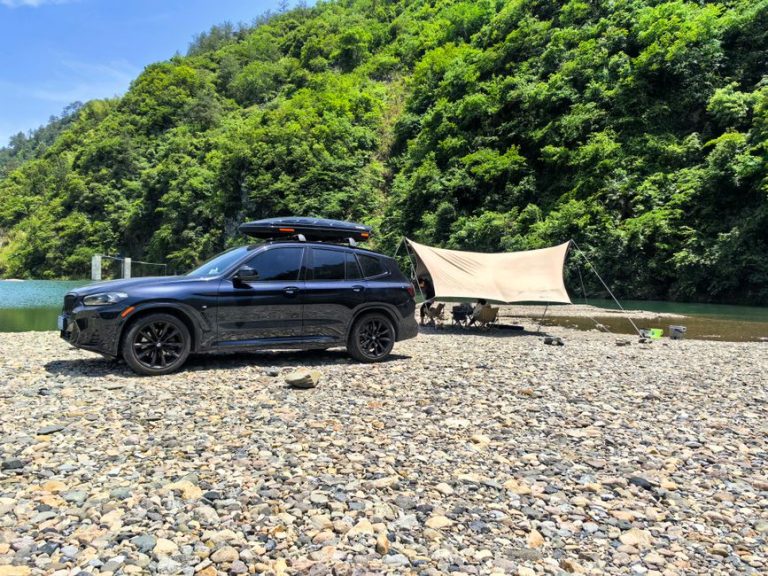 A black SUV parked beside a scenic river with a camping setup under a tent.