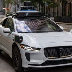 A white autonomous vehicle navigating a city street, reflecting urban architecture in daylight.
