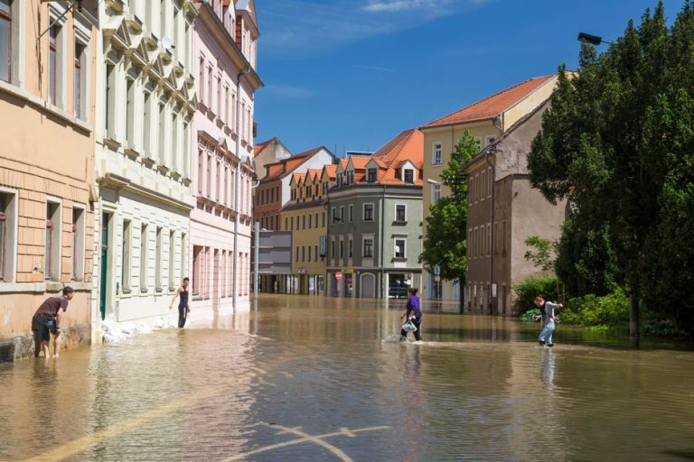 People navigating a flooded street in a European town on a sunny day.