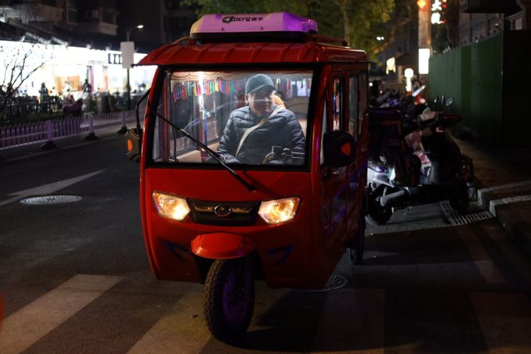 A red tuk-tuk on a busy street at night in Nanjing, China, with a driver inside.