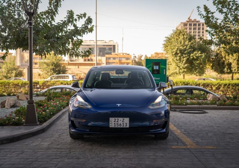 A blue Tesla Model 3 charges at an outdoor station during the day, surrounded by greenery and urban architecture.