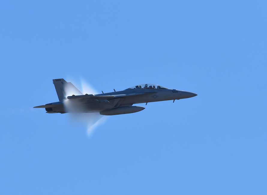 A jet fighter flies at high speed breaking the sound barrier under a clear blue sky.