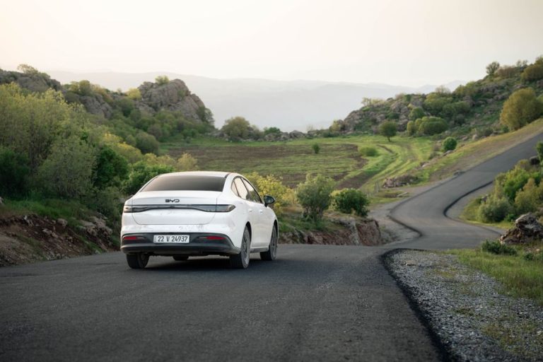 A white car drives on a winding road through scenic hills in Shaqlawa, Iraq during daylight.