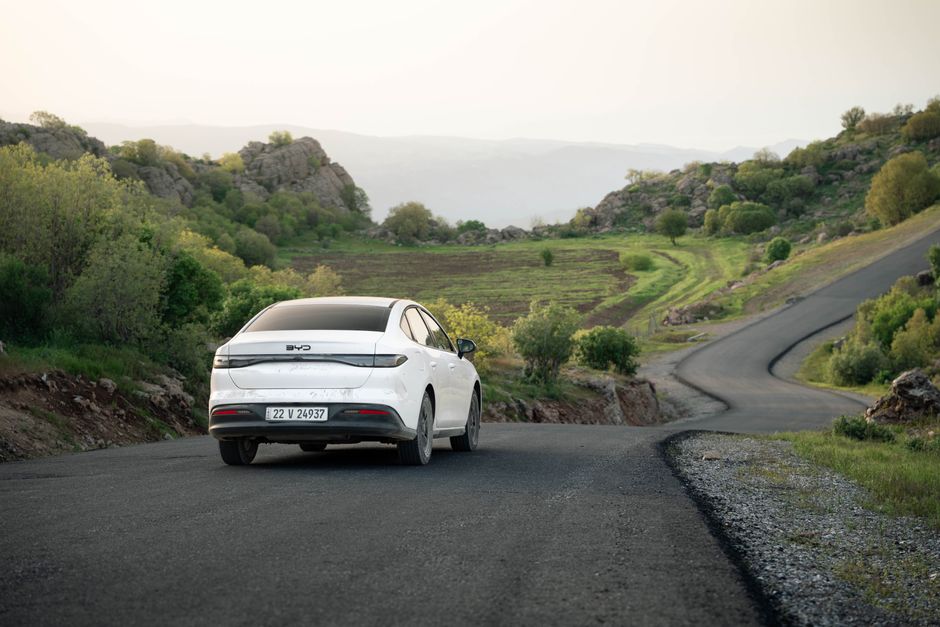 A white car drives on a winding road through scenic hills in Shaqlawa, Iraq during daylight.