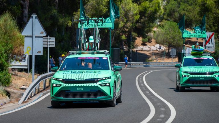 Green electric vehicles with bicycles on roof racks at a cycling race event outdoors.