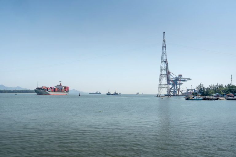 A large container ship sails through an industrial harbor under a clear sky, showcasing maritime logistics.