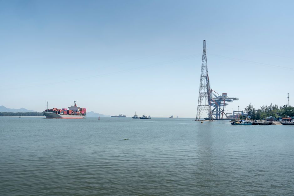 A large container ship sails through an industrial harbor under a clear sky, showcasing maritime logistics.