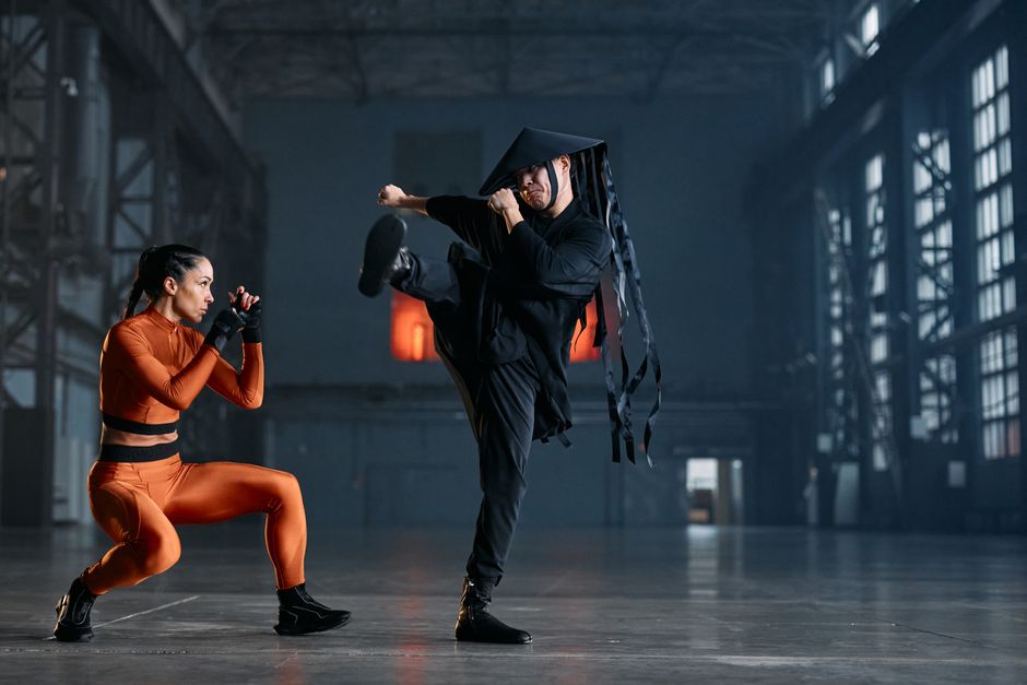 Two cosplayers performing martial arts poses in an industrial space, showcasing vibrant costumes and action.