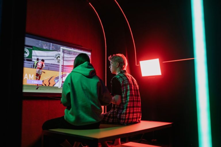 Two friends immersed in gaming on a large screen, surrounded by vibrant RGB lighting in a cozy setup.