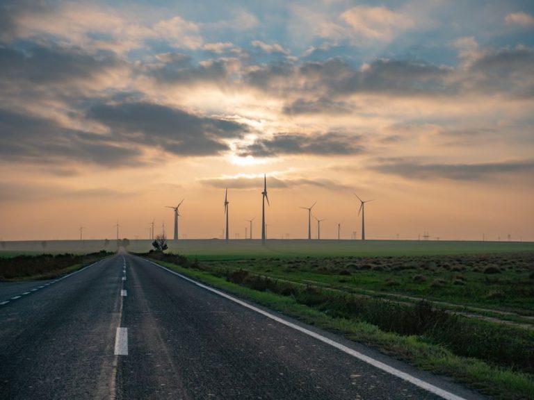 A picturesque rural road leading to wind turbines under a vibrant sunset in Romania, emphasizing renewable energy.