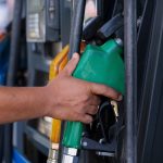 Close-up of a hand holding a green fuel nozzle at a gas pump station outdoors.
