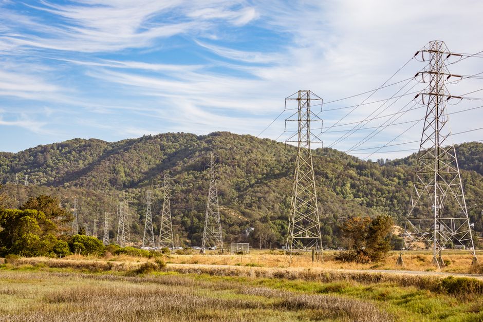 Electric towers dominate a serene countryside landscape with lush mountains under a clear blue sky.