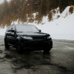 A sleek black SUV navigates a wet, snow-covered mountain road surrounded by evergreen trees.